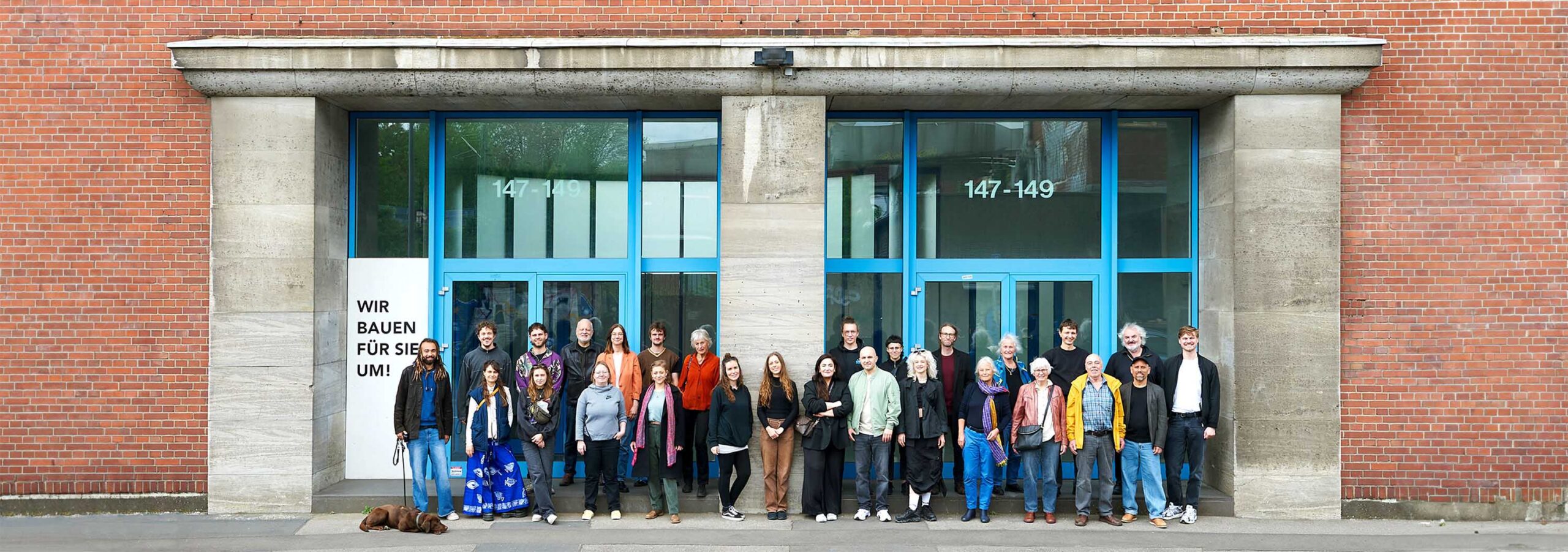 Eine Gruppe von 25 Personen steht vor dem aus blau umrahmten Glastüren bestehenden Haupteingang eines Backsteingebäudes.